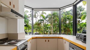 Cozinha com fogão, forno e máquina de lavar louça, janela com vista para palmeiras e micro-ondas no balcão.