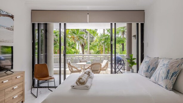 Quarto com cama grande, televisão, cómoda de madeira e varanda com mobiliário de exterior.