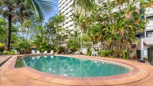 Uma piscina com deck de tijolos, cercada por palmeiras e um edifício alto.