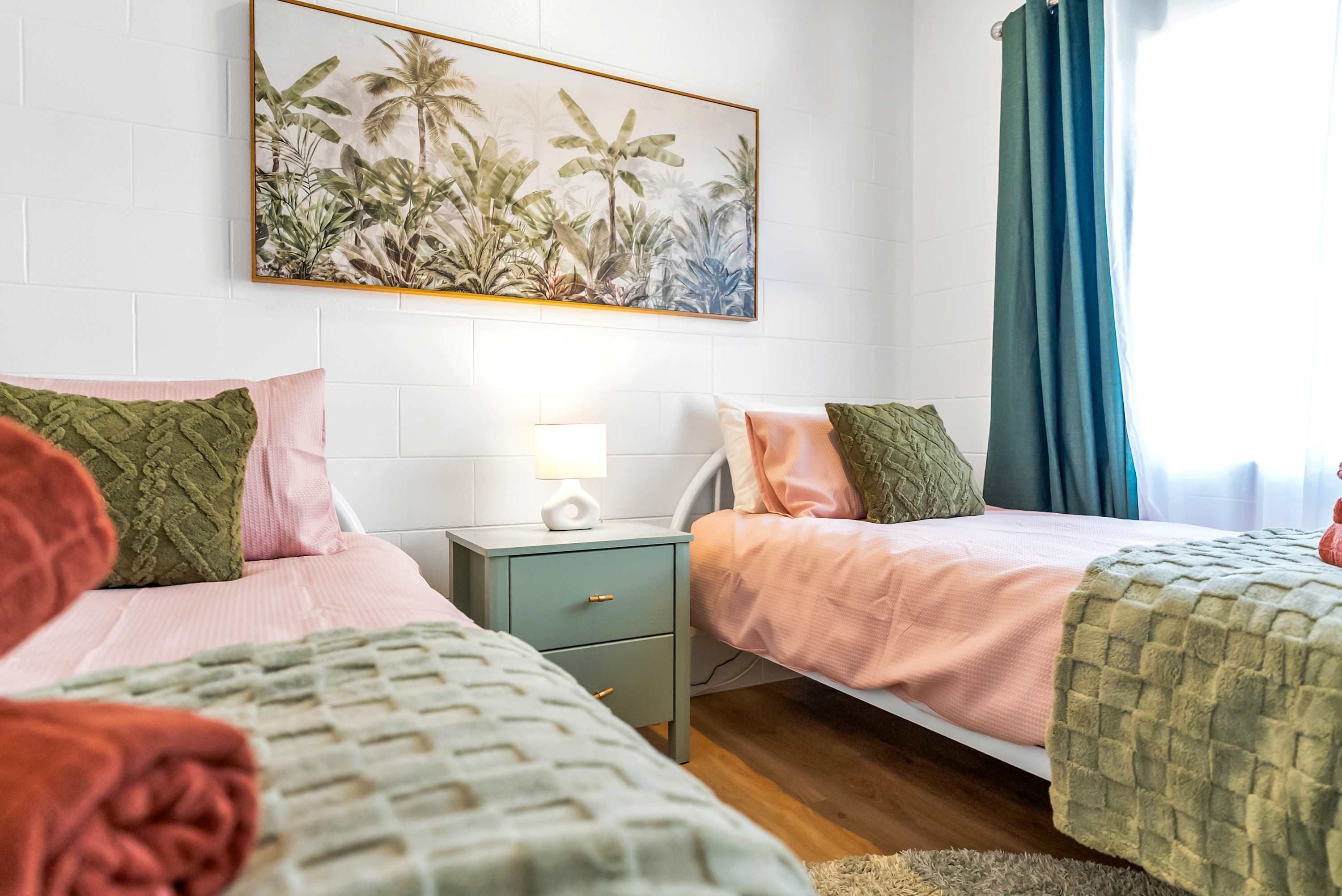 A bedroom with two beds, a nightstand, a lamp, and a tropical-themed wall art.