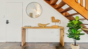 Een houten console tafel met een spiegel, een houten sculptuur en een potplant in een kamer met een stenen vloer en een houten trap.