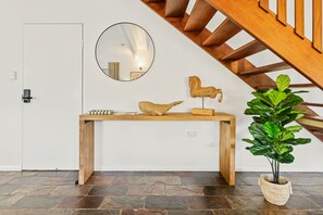 A wooden console table with a mirror, a wooden sculpture, and a potted plant in a room with a stone floor and a wooden staircase. - Spacious Whitfield Townhouse w Private Garden BBQ (Cairns)