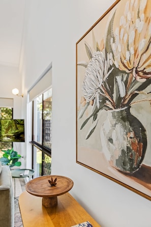 A room with a wooden coffee table, a framed painting of flowers, and a view of greenery outside. - Spacious Whitfield Townhouse w Private Garden BBQ (Cairns)
