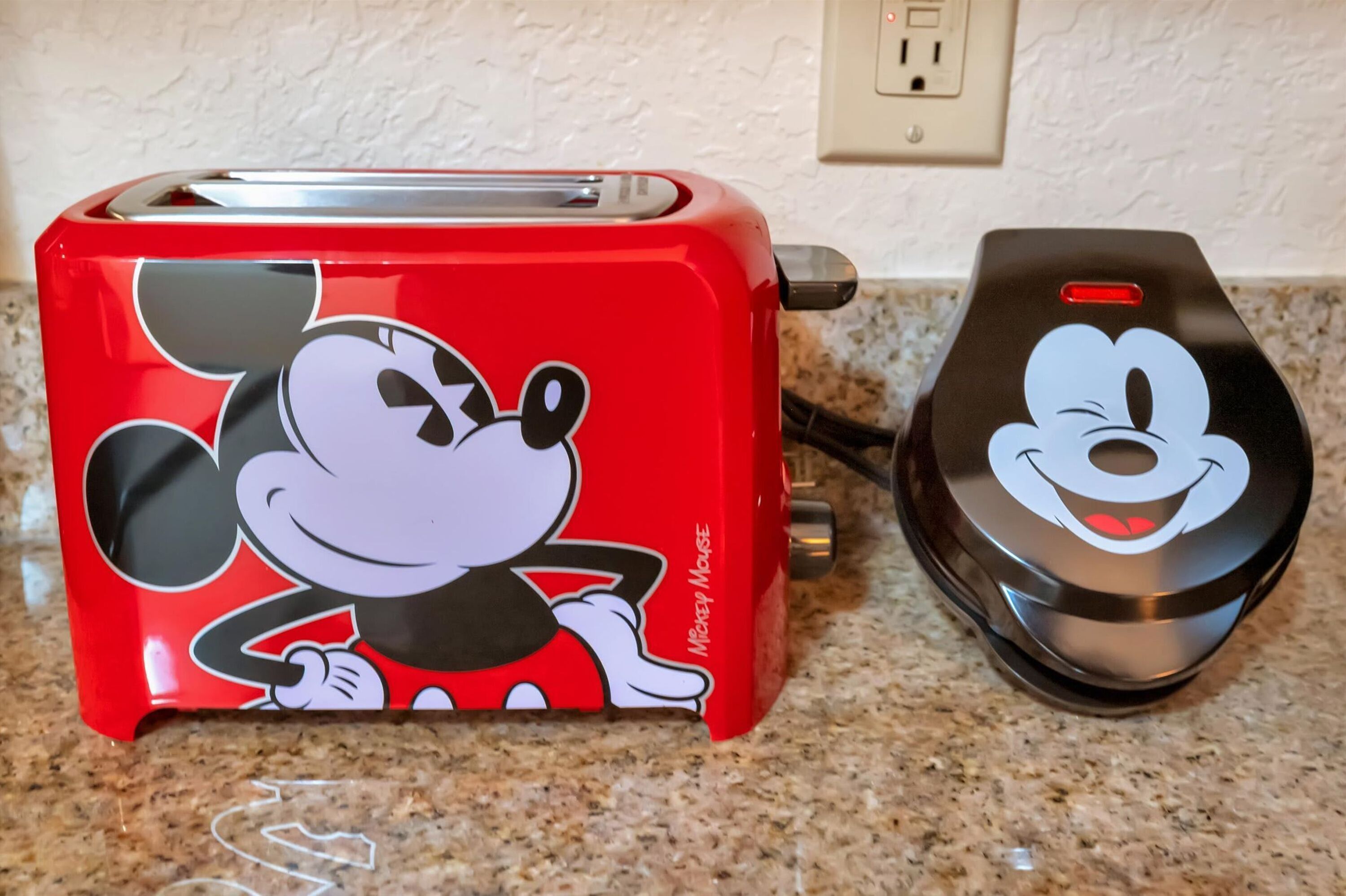 A red toaster with a Mickey Mouse design and a black mouse-shaped device on a kitchen counter.