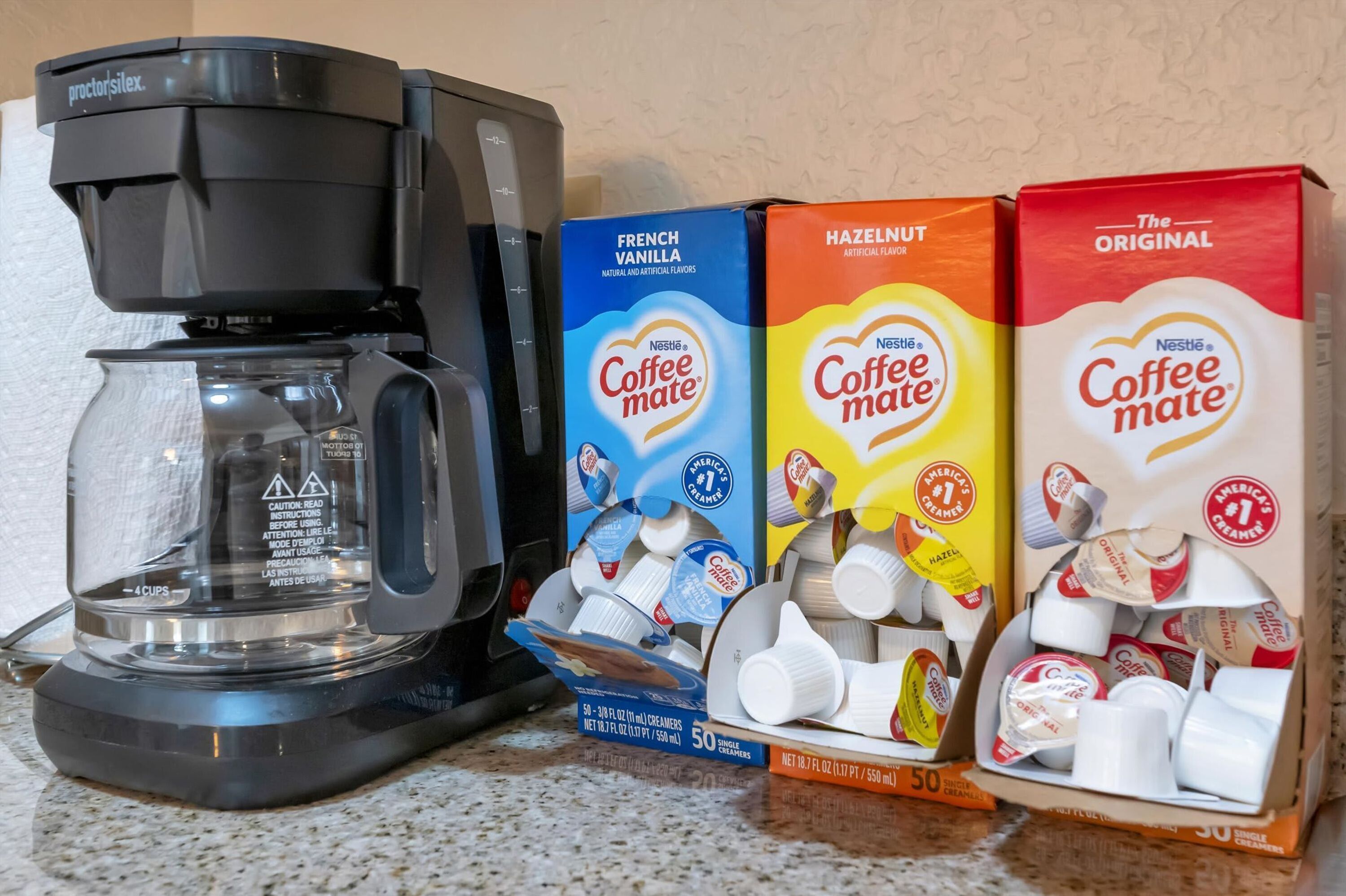 Een koffiezetapparaat, drie pakken Nestle Coffee Mate melkpoeder en een glazen kan op een aanrecht.