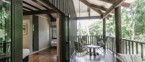Una terraza de madera con una hamaca colgante y una mesa, una habitación con cama y alfombra, y vistas al verdor exterior.
