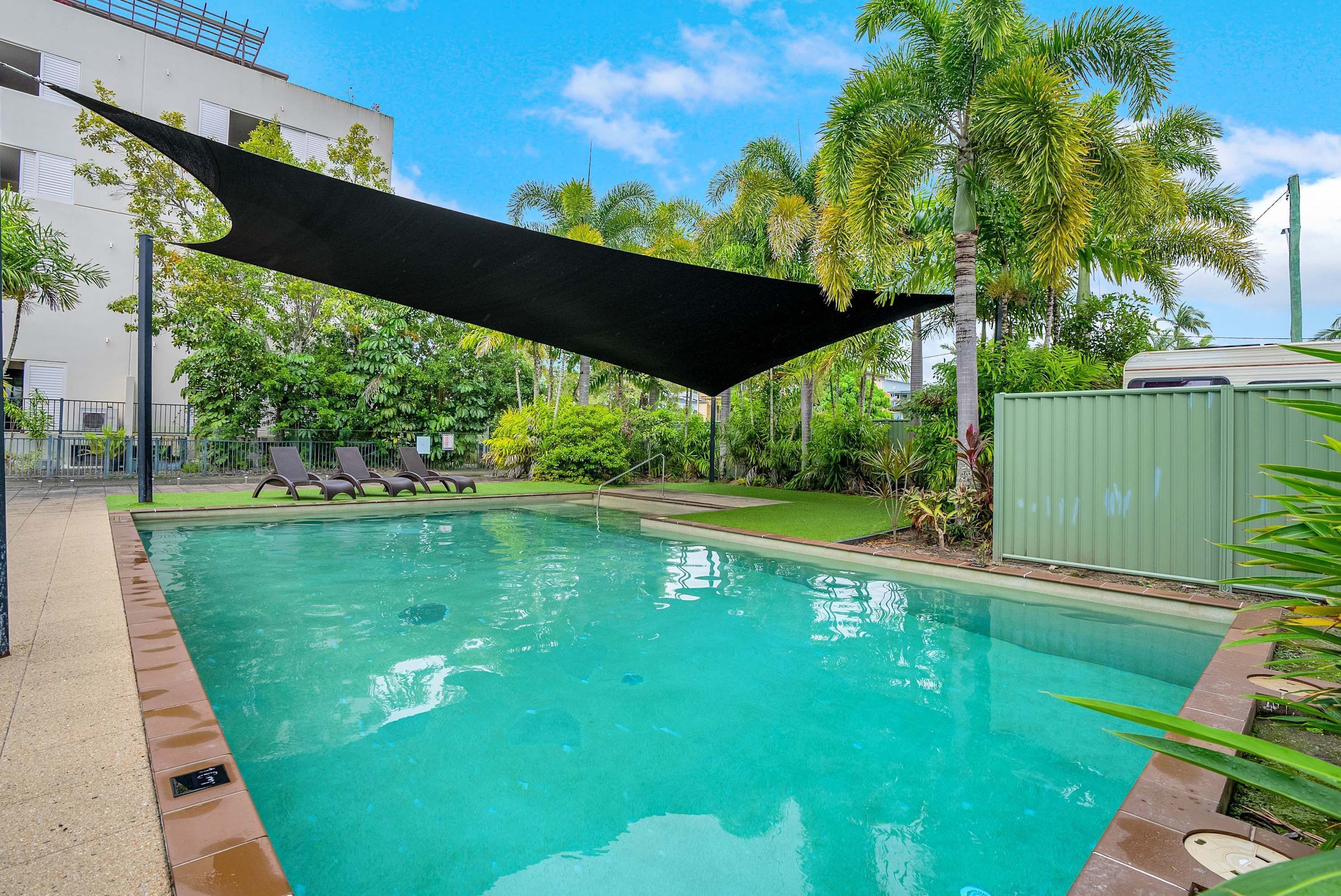 A swimming pool with a black shade sail, surrounded by palm trees and a building in the background.