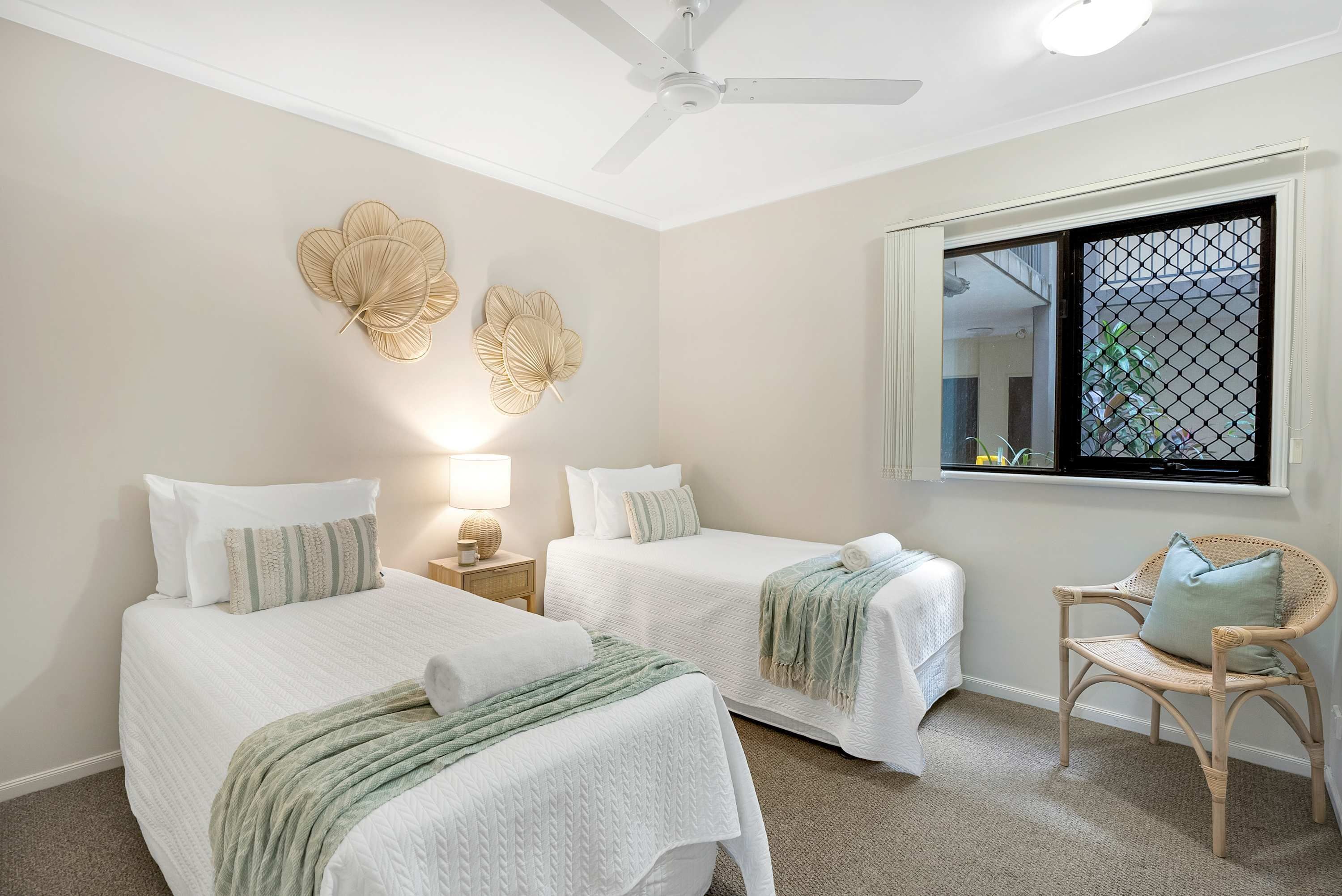 A bedroom with two beds, a rattan chair, a ceiling fan, and wall art.