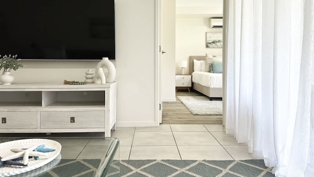 A modern living room with a flat-screen TV, a white entertainment unit, and a dining area with a glass table. - The Nook - Pet Family-friendly 4-bedroom (Cairns)