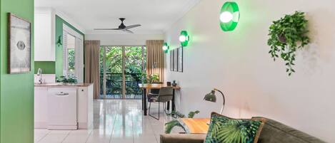 A modern kitchen with white cabinets, a dining area with a table and chairs, a sofa with a tropical pillow, and a balcony with greenery.