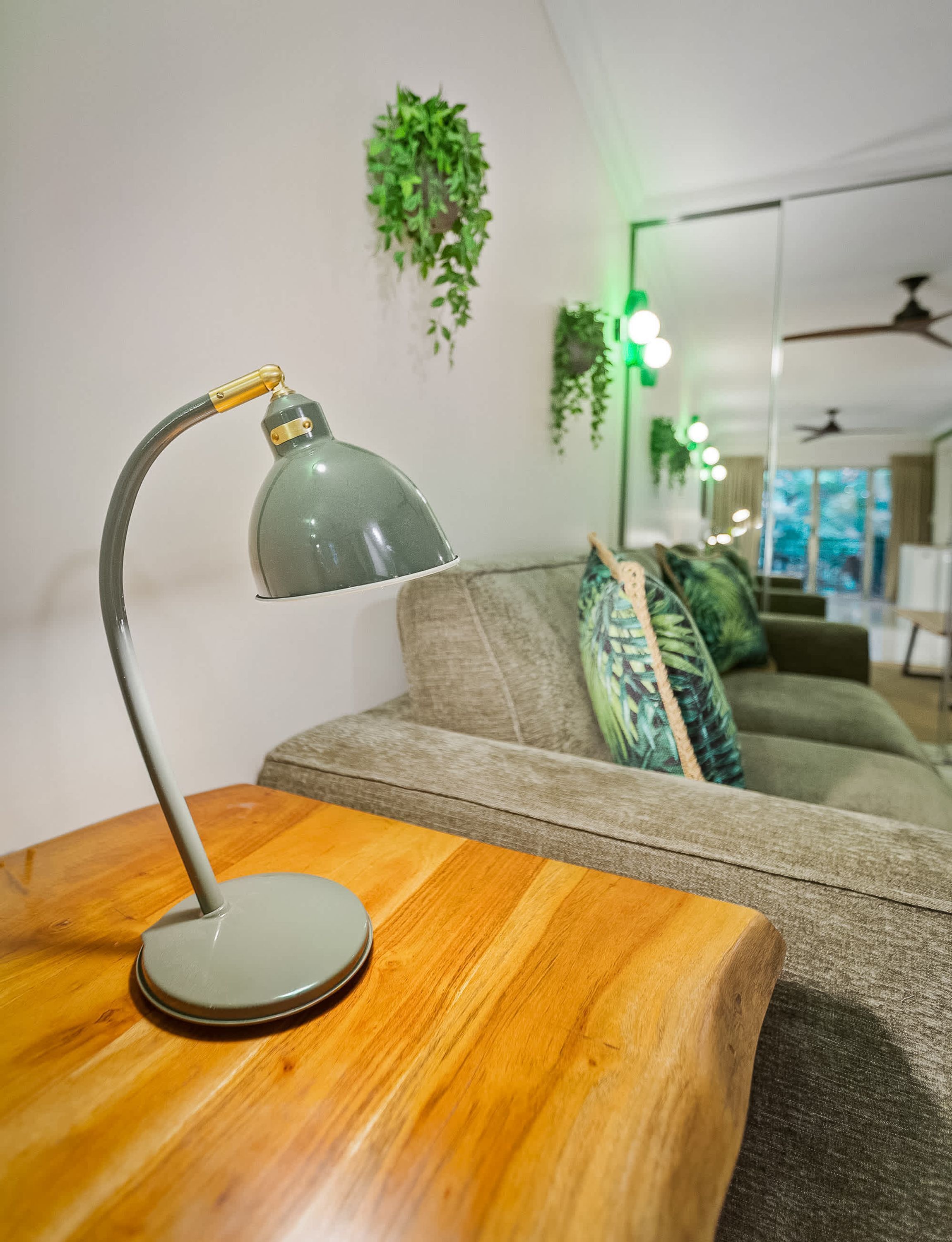 Un salon moderne avec une table d’appoint en bois, une lampe de bureau grise, un canapé gris et des plantes vertes accrochées au mur.