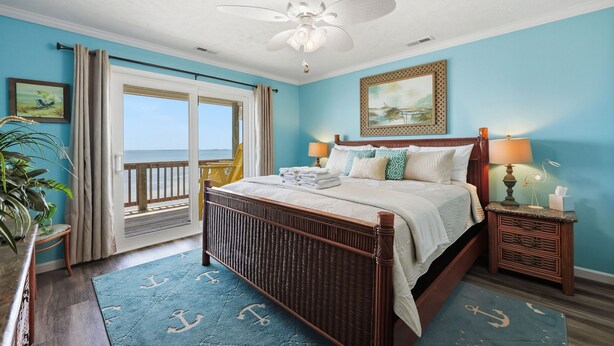 Een slaapkamer met een bed, nachtkastje, plafondventilator, raam met gordijnen en uitzicht op zee.