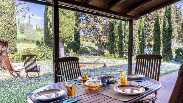 Ein Essbereich im Freien mit einem für zwei Personen gedeckten Tisch, umgeben von Stühlen und Blick auf einen Garten mit Bäumen und einer hölzernen Struktur im Hintergrund.