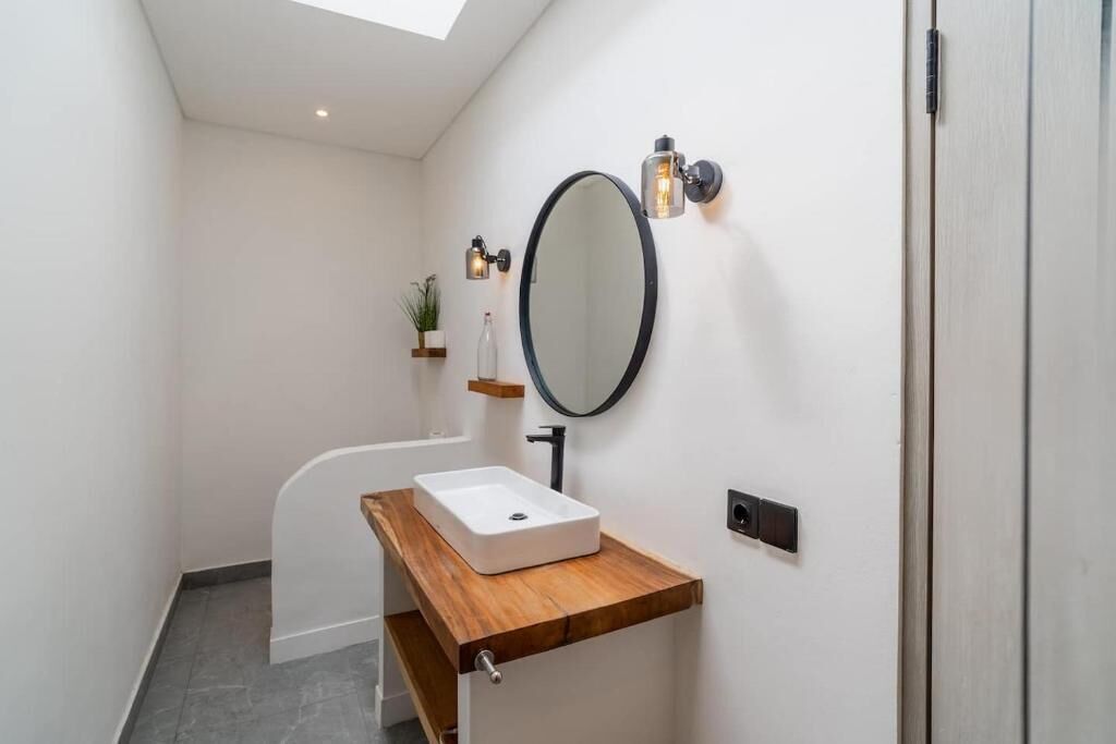 Une salle de bain avec un lavabo blanc, un plan de toilette en bois, un miroir rond et deux appliques murales.