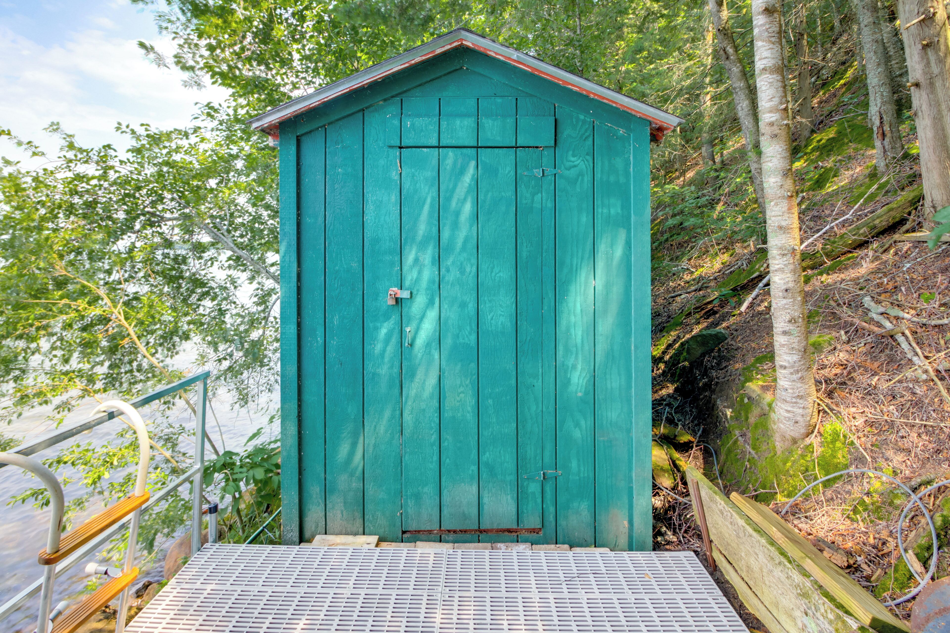 A green wooden shed with a metal staircase leading up to it, surrounded by trees and a body of water.