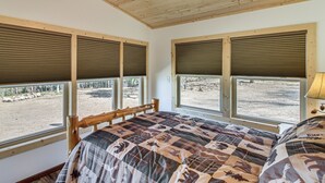 Une chambre avec un plafond en bois, quatre fenêtres équipées de stores, un lit recouvert d’une couvre-lit à motifs et une vue sur un paysage naturel.