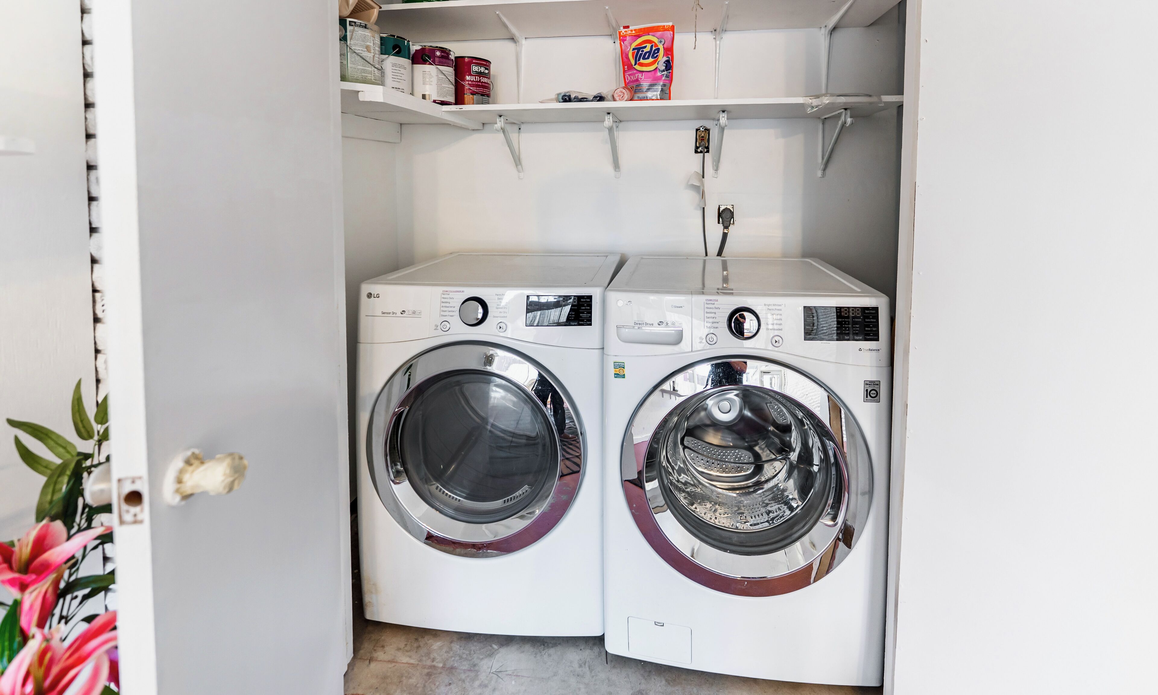 A laundry room with two white washing machines, a shelf with cleaning supplies, and a door with a floral design.