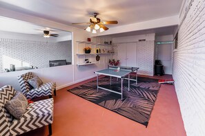 A modern living room with a ping pong table, a sofa with patterned cushions, a ceiling fan, and a brick-patterned wall. - Large Deck, Rec Room! 2-unit ATX Retreat By Campus (Austin)
