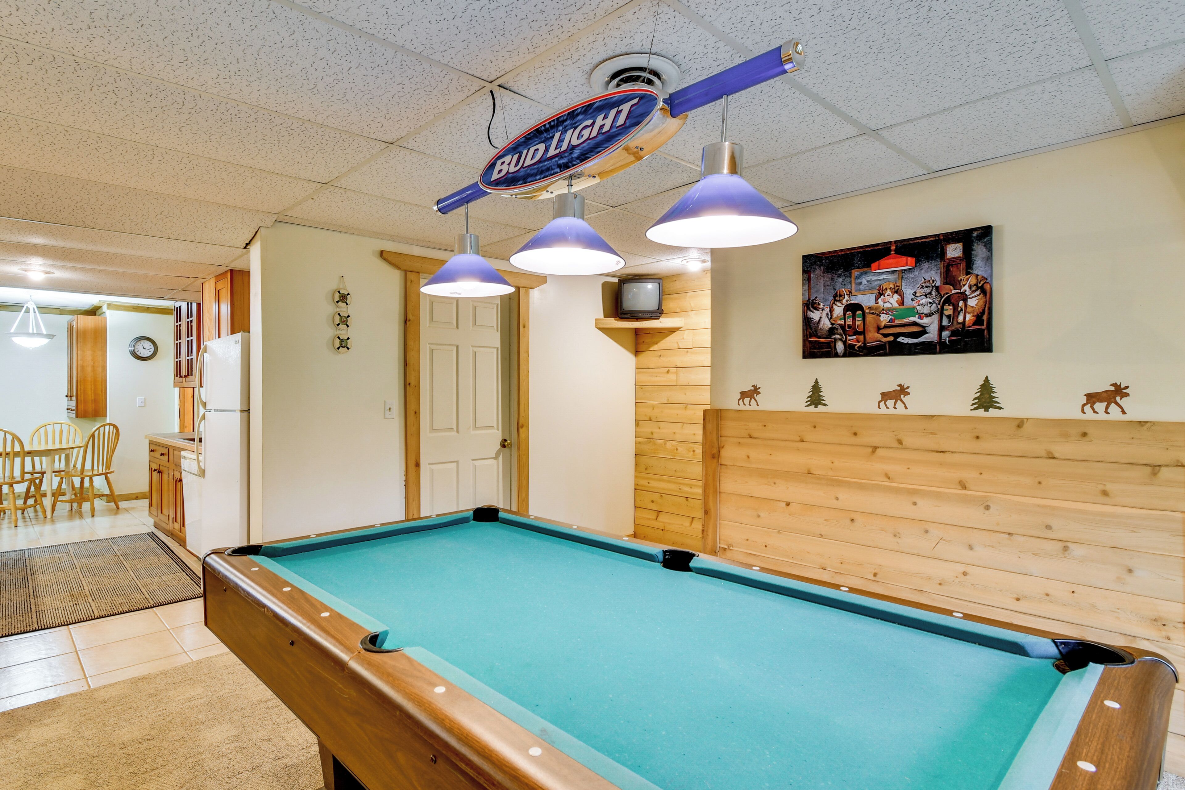 Une table de billard éclairée par des lampes suspendues, un mur en bois orné d’une fresque et une enseigne publicitaire pour la bière Bud Light.