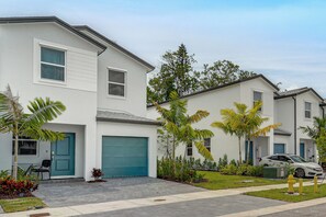 A modern two-story residential building with a garage, a driveway, and a well-maintained lawn with palm trees.