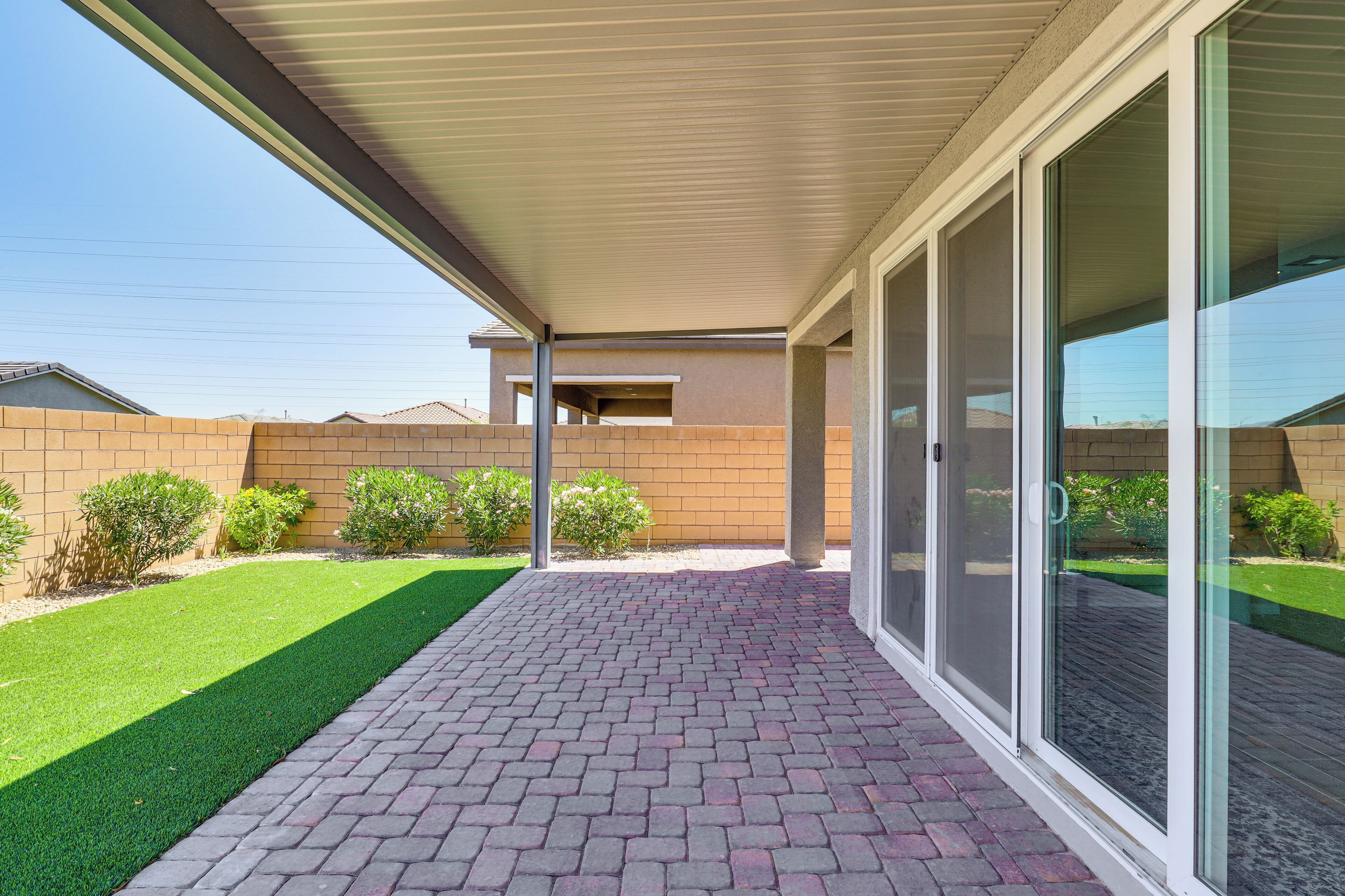 A covered patio with a brick floor, glass sliding doors, and a view of a backyard with a lawn and bushes.