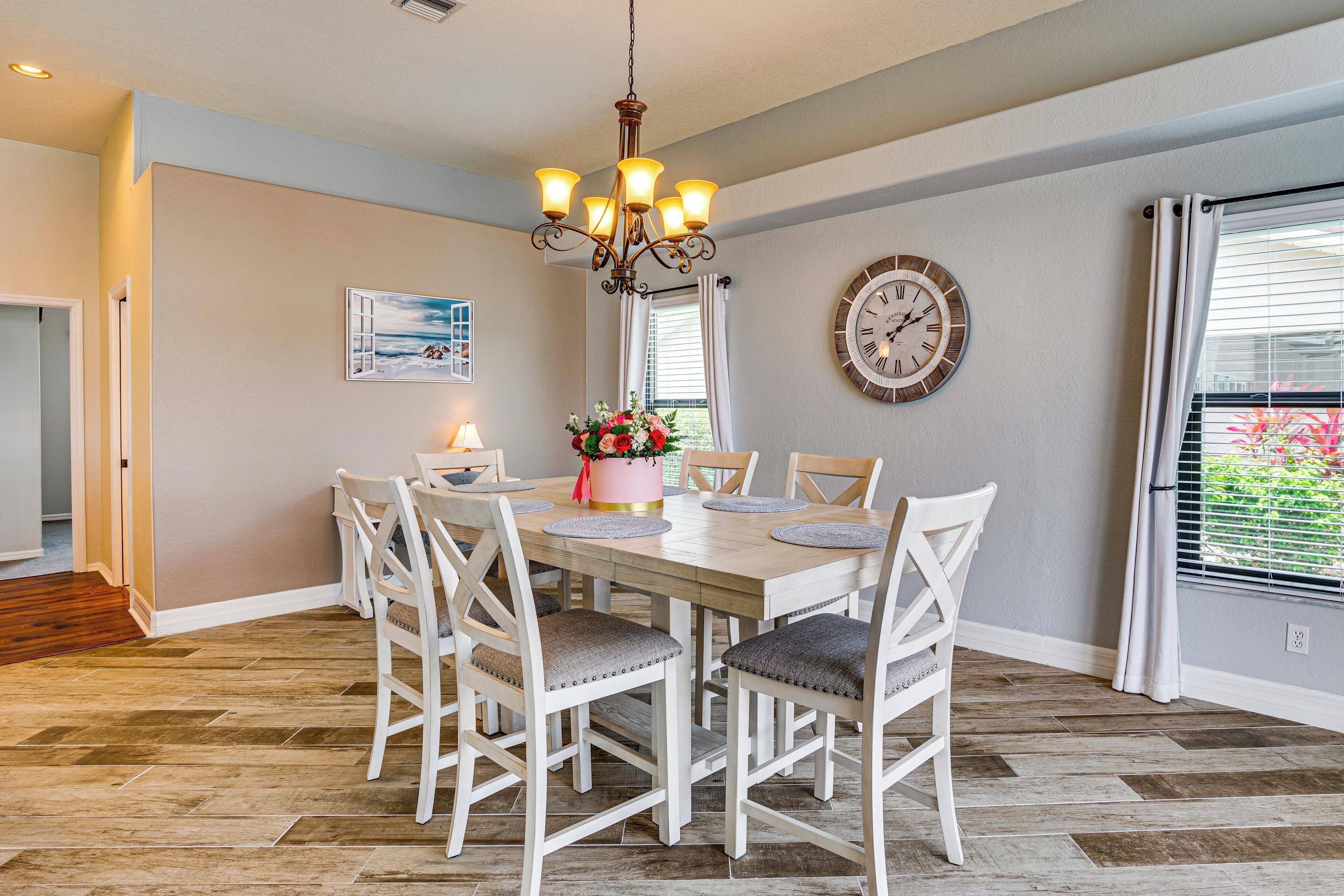 Une salle à manger avec une table en bois, des chaises blanches, un lustre, une horloge et une fenêtre avec des stores.