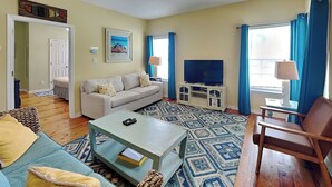 Un salon avec un ventilateur de plafond, un canapé, une table basse, un téléviseur posé sur un meuble et un tapis à motifs.