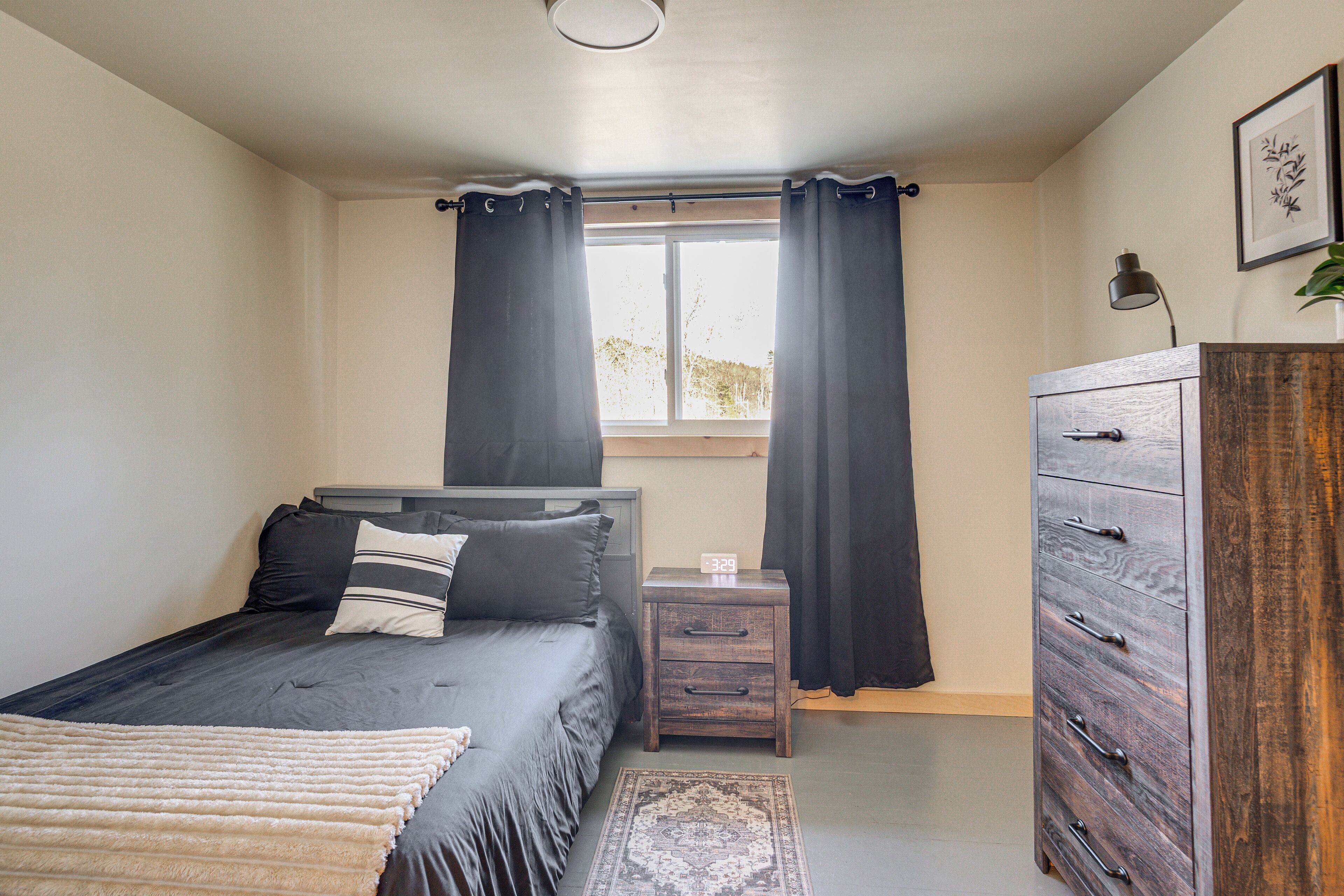 A bedroom with a bed, a nightstand, a dresser, a window with curtains, and a small rug.