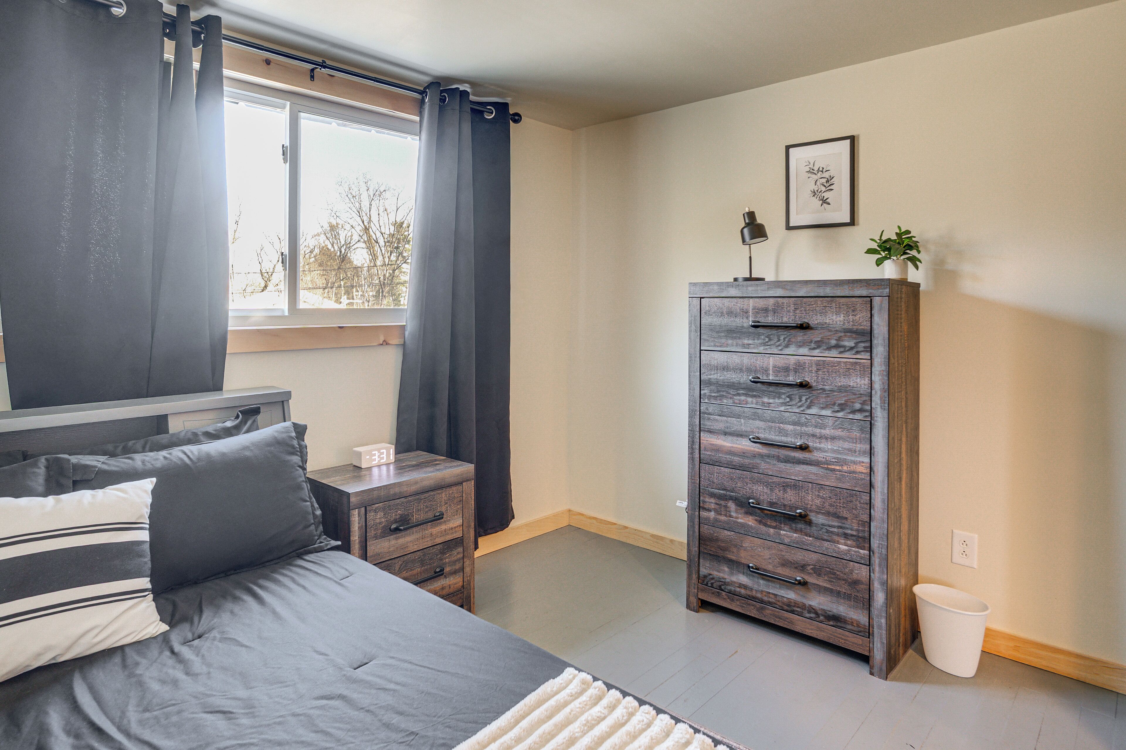 Quarto com cama, cômoda, mesa de cabeceira, janela com cortinas, planta presa na parede e um quadro emoldurado.
