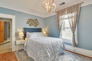 A bedroom with a bed, nightstands, a chandelier, a window with curtains, and a decorative wall piece.