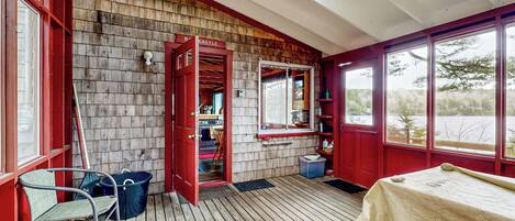 Um deck de madeira com um banco, uma mesa e uma porta vermelha que dá para um quarto com janela e prateleira.