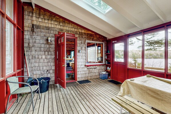 A wooden deck with a bench, a table, and a red door leading to a room with a window and a shelf. - Peaceful 2BR Lakefront Cabin w/ New Dock (Mount Desert)