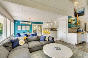 A modern living room with a sectional sofa, a round coffee table, and a kitchen area with white cabinets and a wooden staircase. - Lakefront Bliss w/ Dock + Fire Pit! Ozarks Home (Osage Beach)