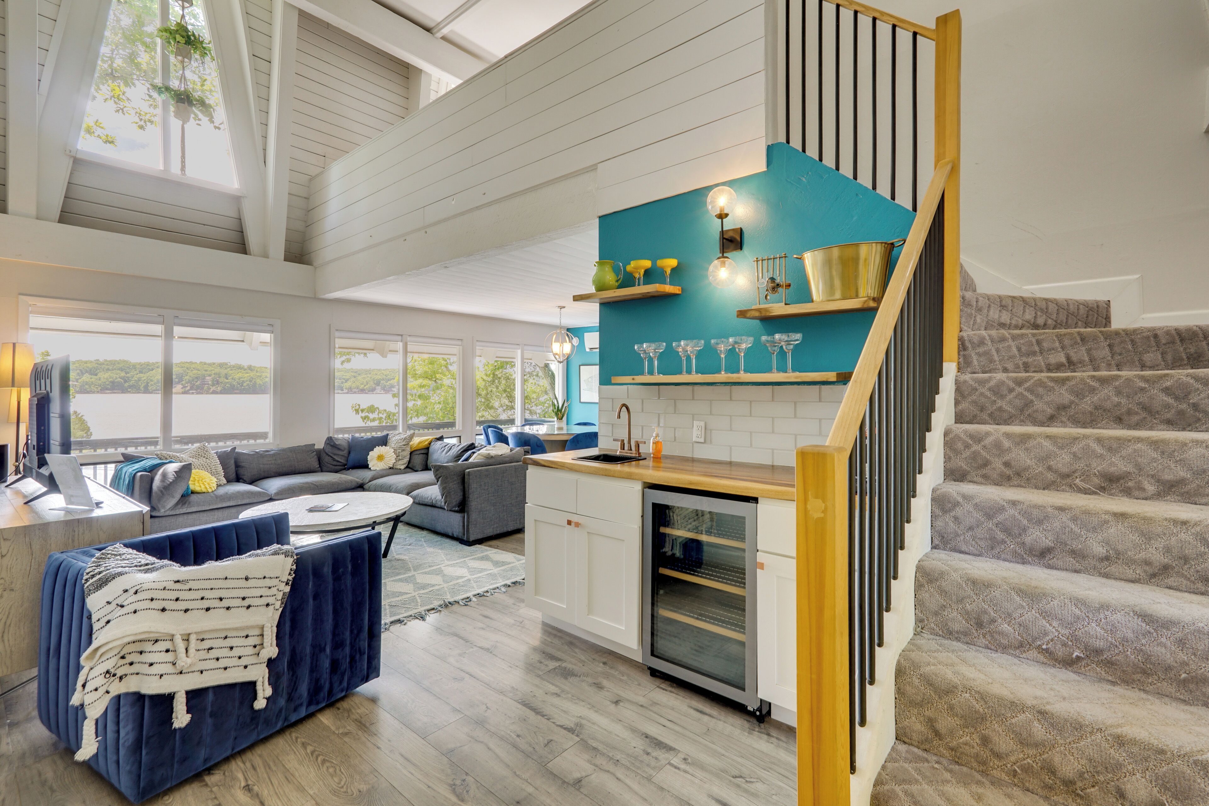 A spacious living room with a sofa, a blue ottoman, and a kitchen with a blue accent wall, a staircase, and a view of the outdoors.