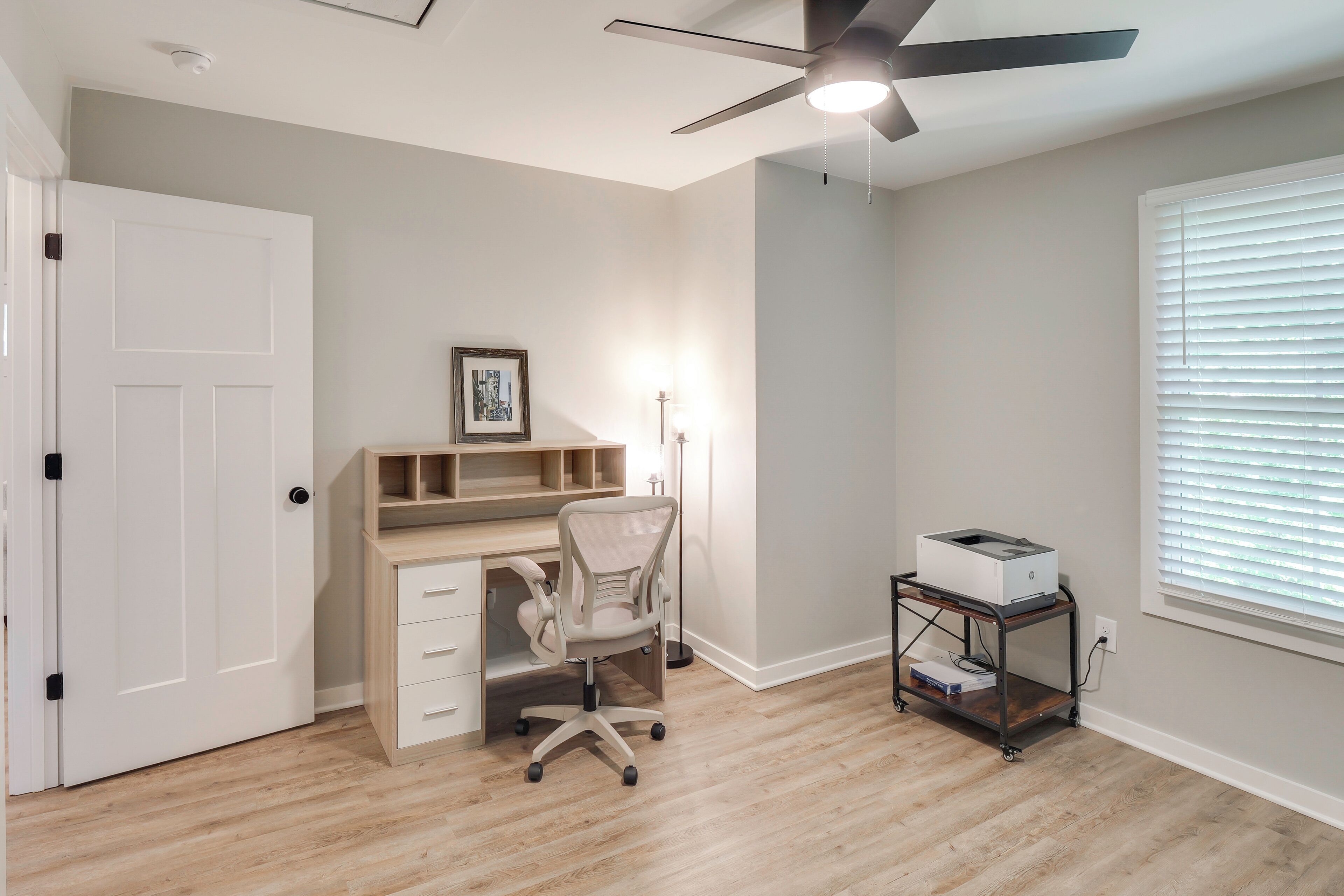A room with a ceiling fan, a desk with drawers, a chair, a printer on a cart, and a window with blinds.