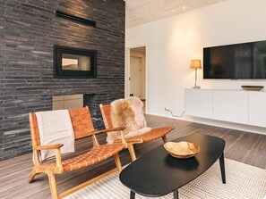Un salon moderne avec un mur en briques, deux fauteuils en bois, une table basse noire, un téléviseur et une cheminée.