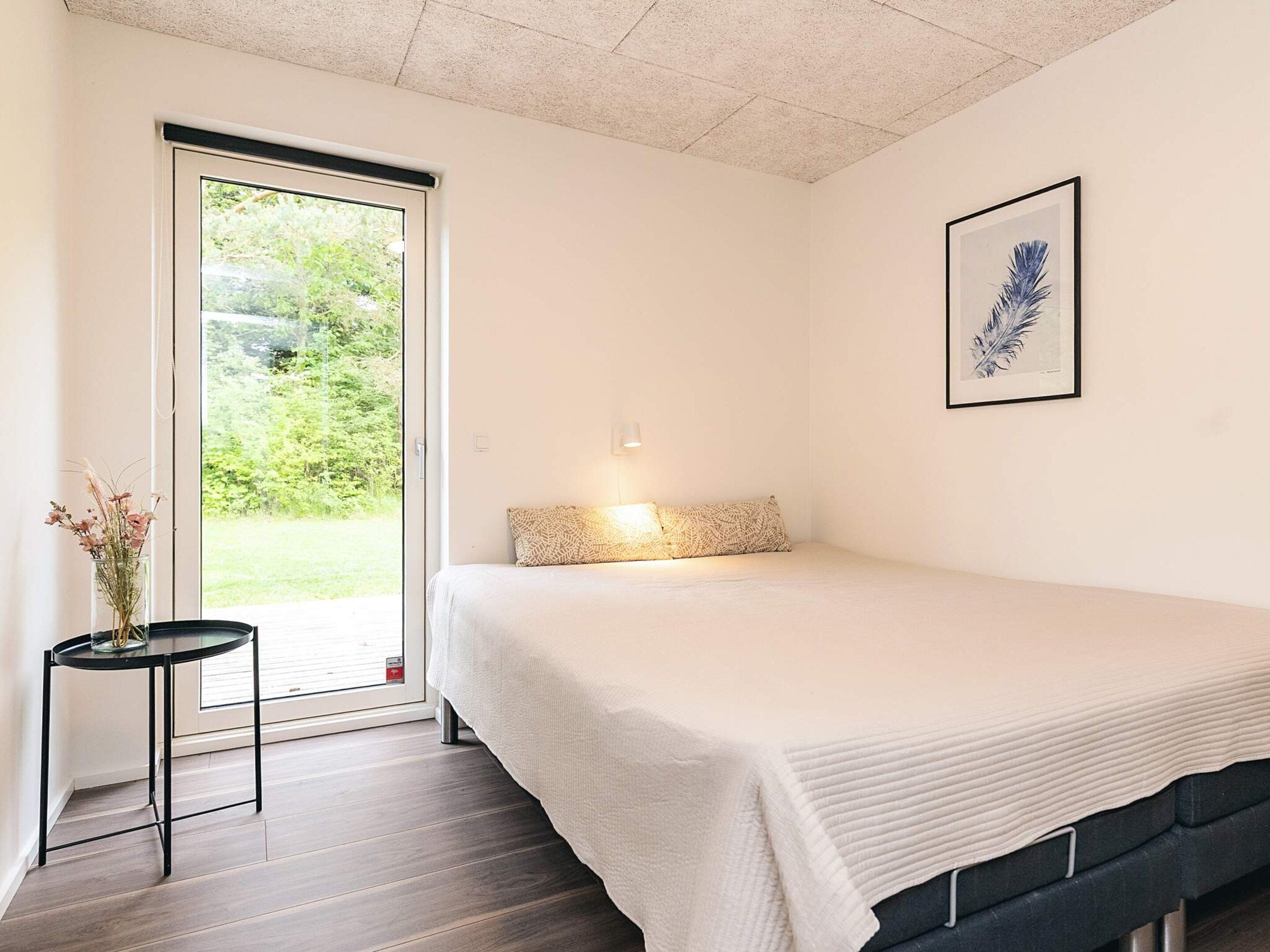 Un dormitorio con cama, una mesita de noche con un florero, un cuadro en la pared y vistas por la puerta.