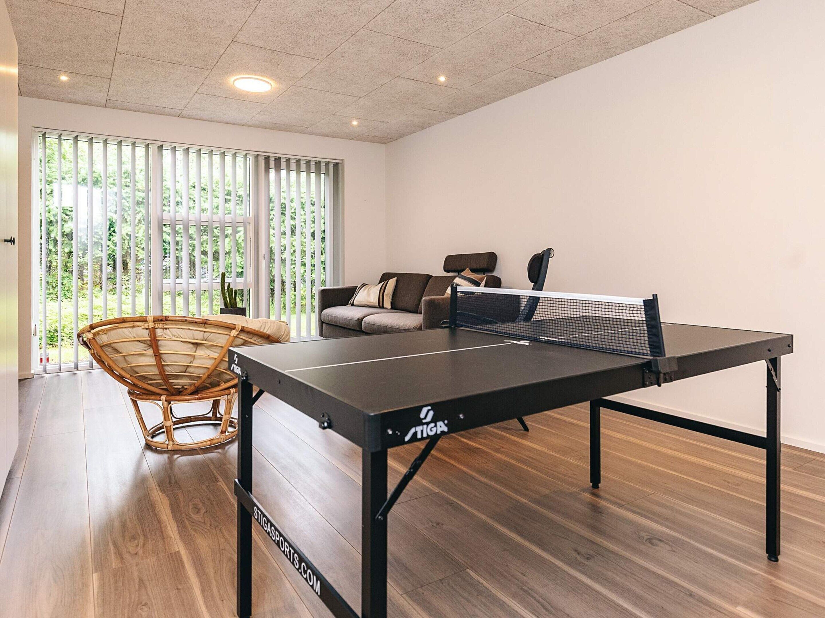 A ping pong table in a room with a wooden floor, a wicker chair, a sofa, and a window with vertical blinds.