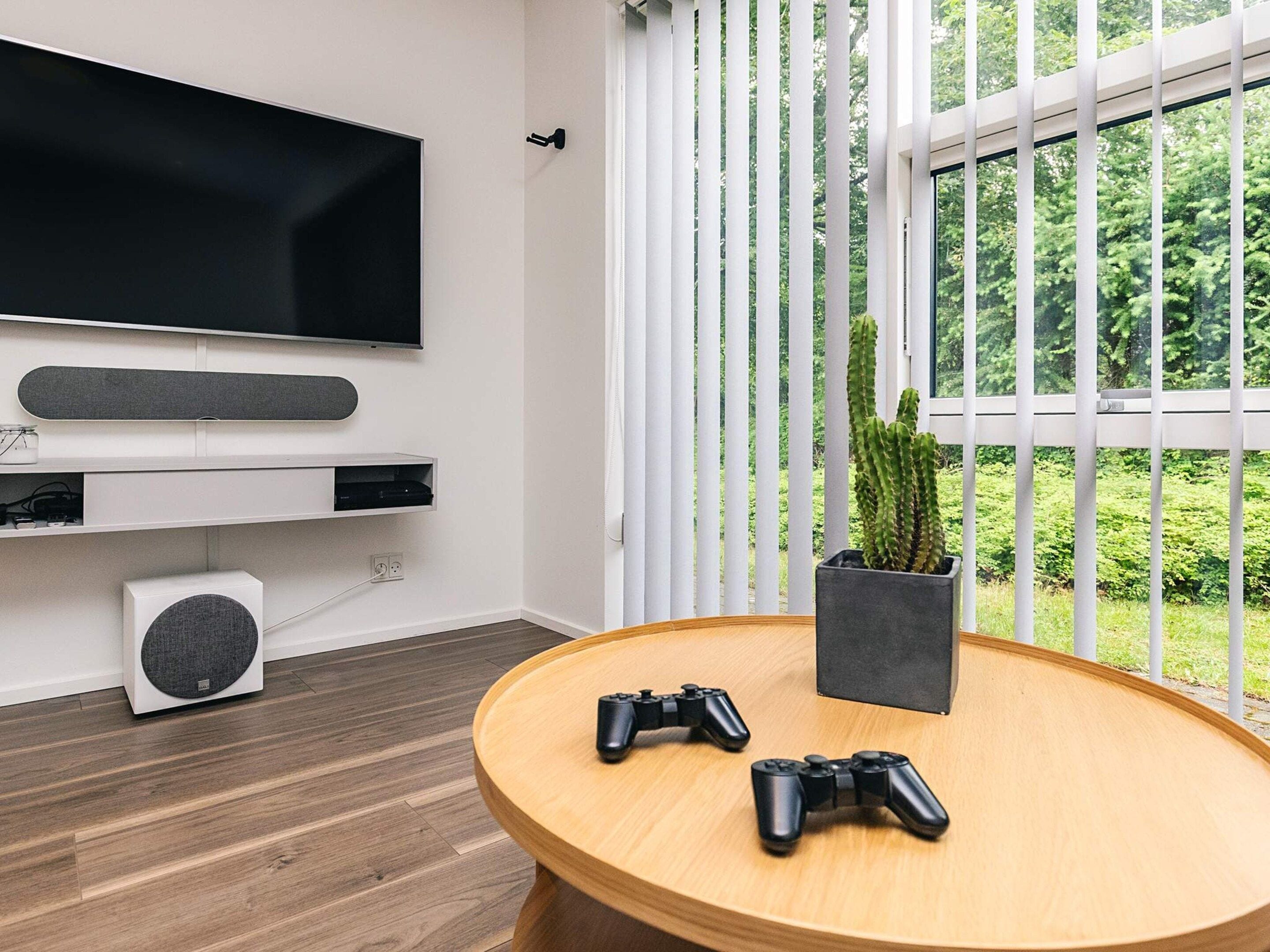 Una sala moderna con un televisor de pantalla plana montado en la pared, una mesa redonda de madera con dos controles de videojuegos, una planta en maceta y una ventana con persianas verticales.
