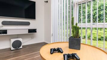 Una sala moderna con un televisor de pantalla plana montado en la pared, una mesa redonda de madera con dos controles de videojuegos, una planta en maceta y una ventana con persianas verticales.