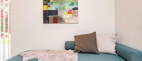 A teal sofa with two pillows and a fur throw, a colorful abstract painting on the wall, and a window with blinds partially visible.