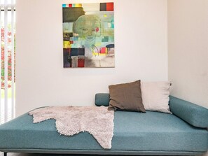 Ein blaugrünes Sofa mit zwei Kissen und einer Felldecke, ein farbenfrohes abstraktes Gemälde an der Wand und ein Fenster mit teilweise sichtbaren Jalousien.