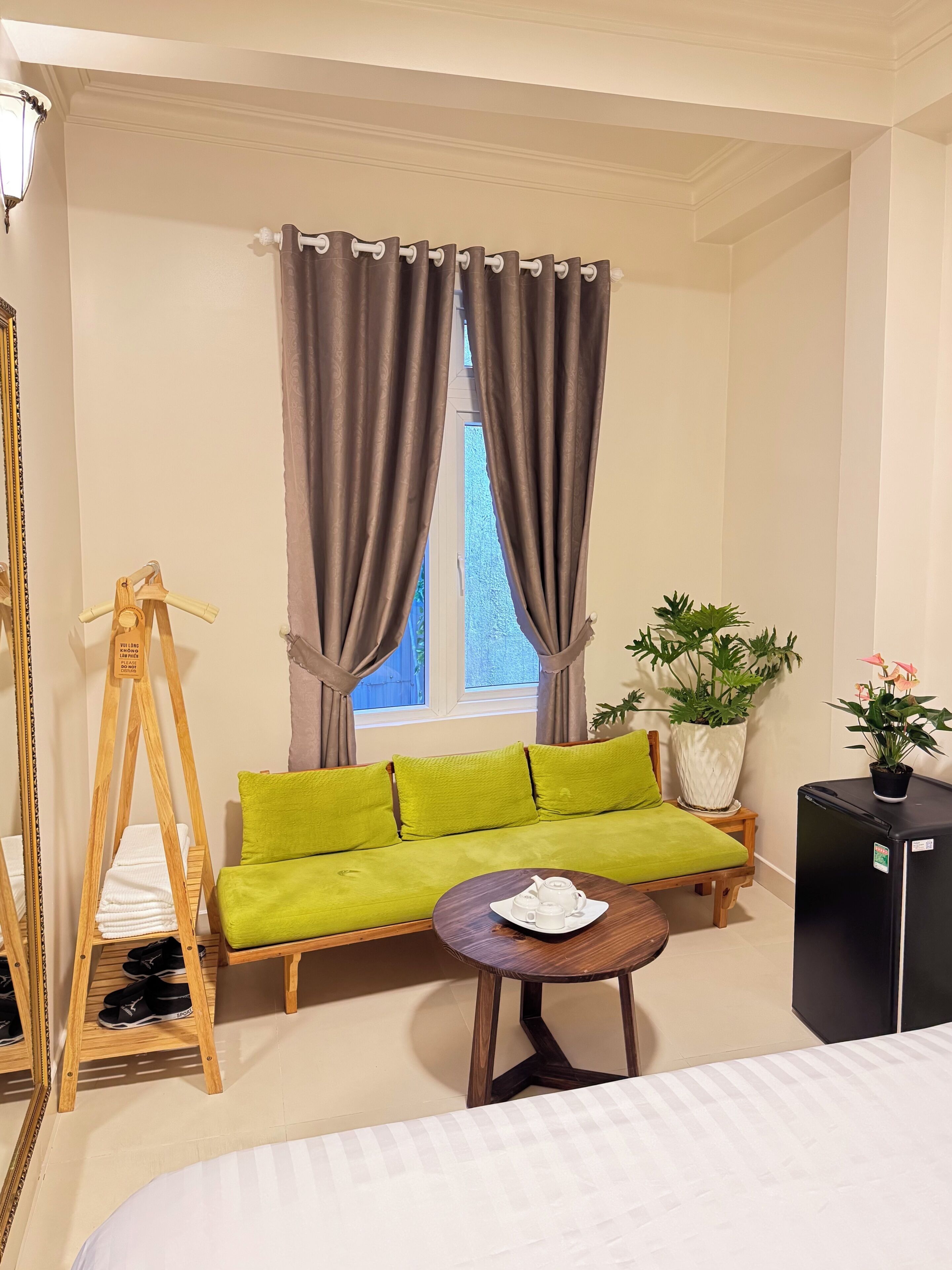 A room with a green sofa, a small round table with a teapot, a wooden shoe rack, a potted plant, and a black refrigerator.