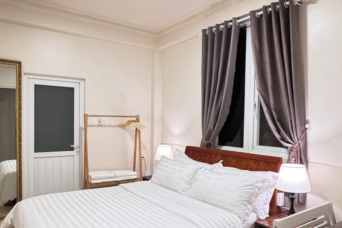A hotel room with a bed, a wooden nightstand, a table, a mirror, and a window with curtains.