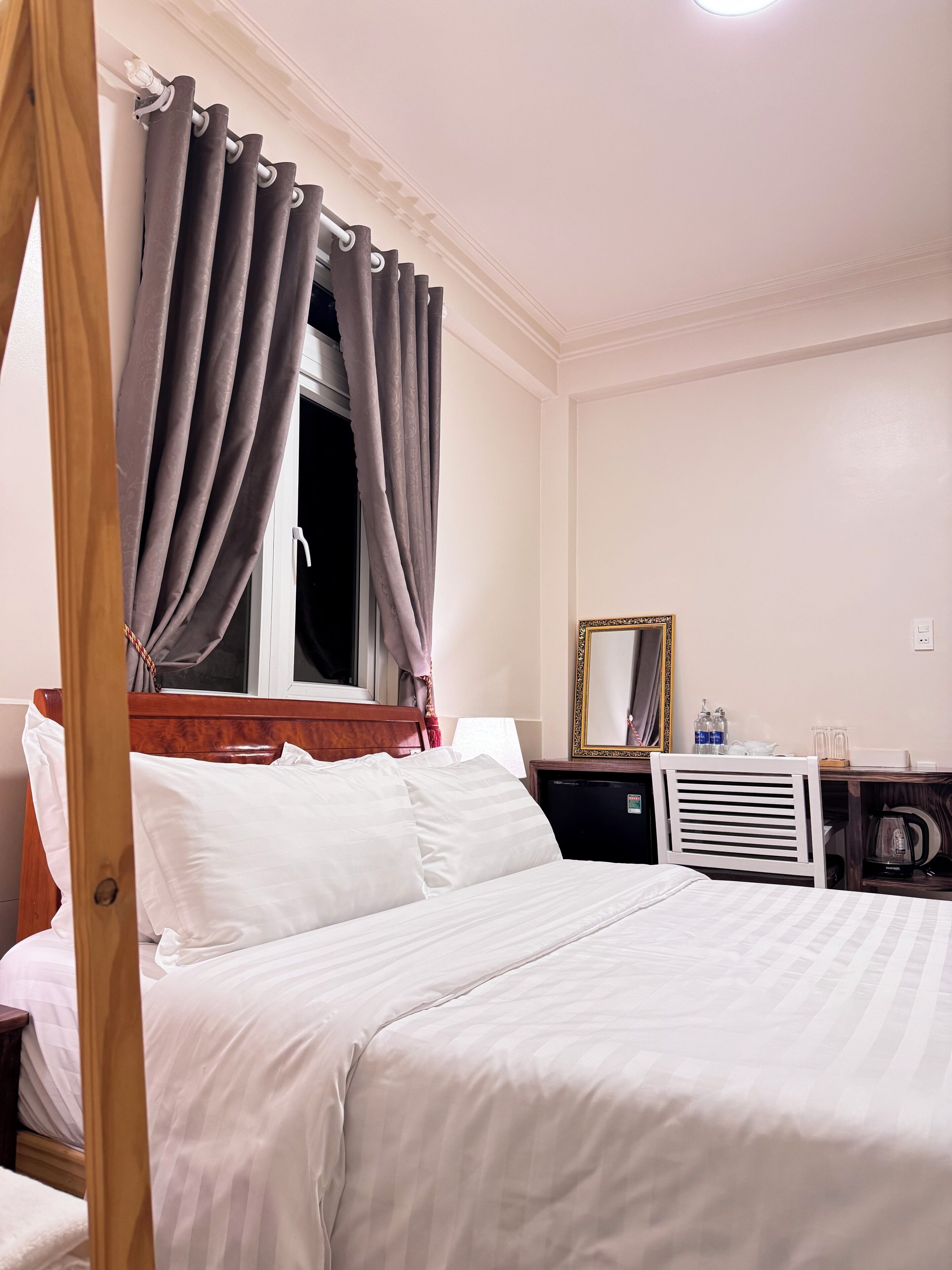 A hotel room with a bed, a wooden headboard, a nightstand, a dresser, a mirror, and a window with curtains.