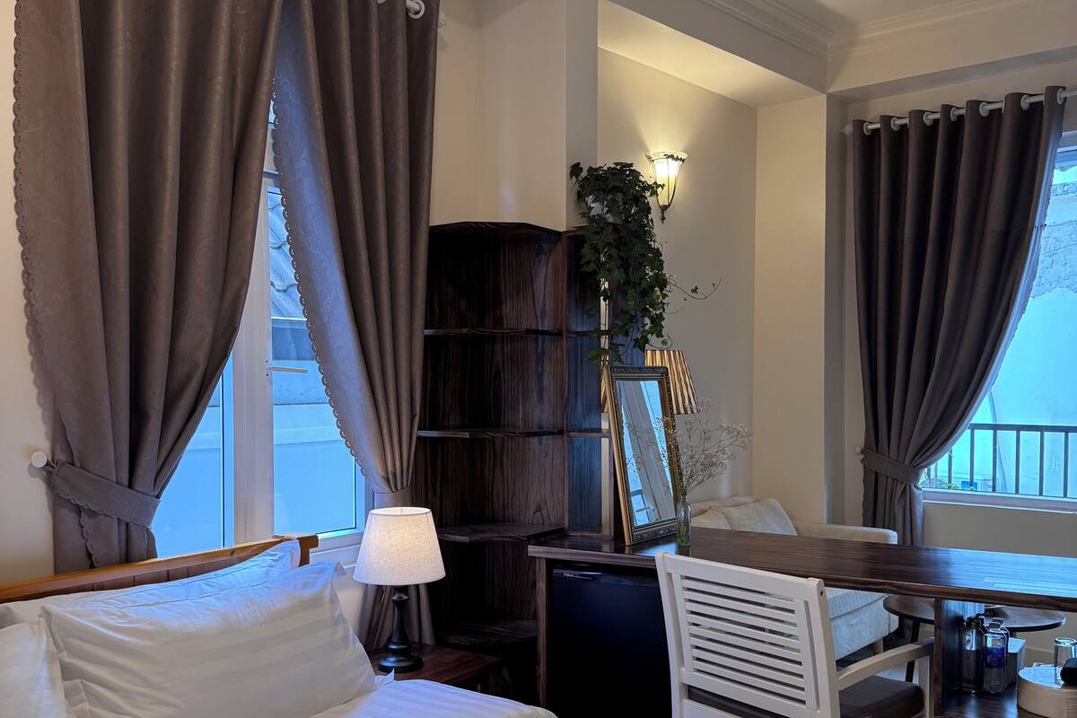 A hotel room with a bed, a desk, a chair, a lamp, a bookshelf, and a window with curtains.