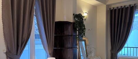 A hotel room with a bed, a desk, a chair, a lamp, a bookshelf, and a window with curtains.