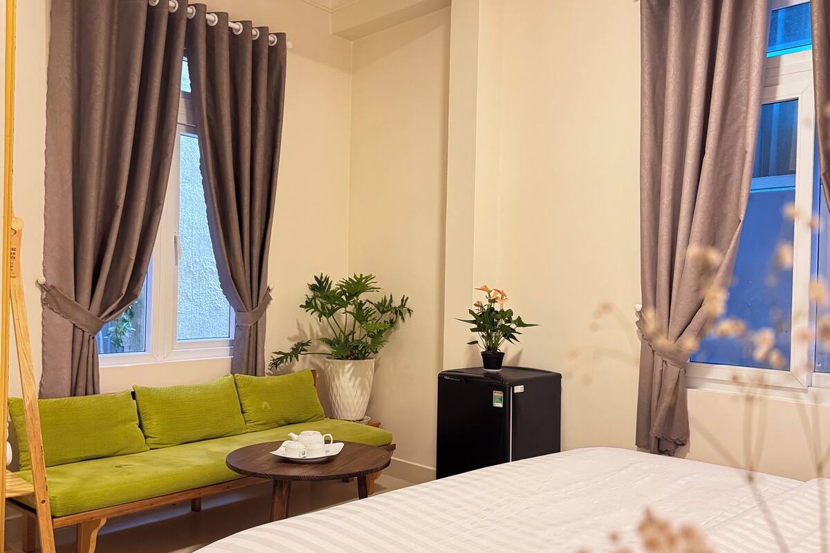 A hotel room with a bed, a green sofa, a small table with a tray, a potted plant, and a black refrigerator.