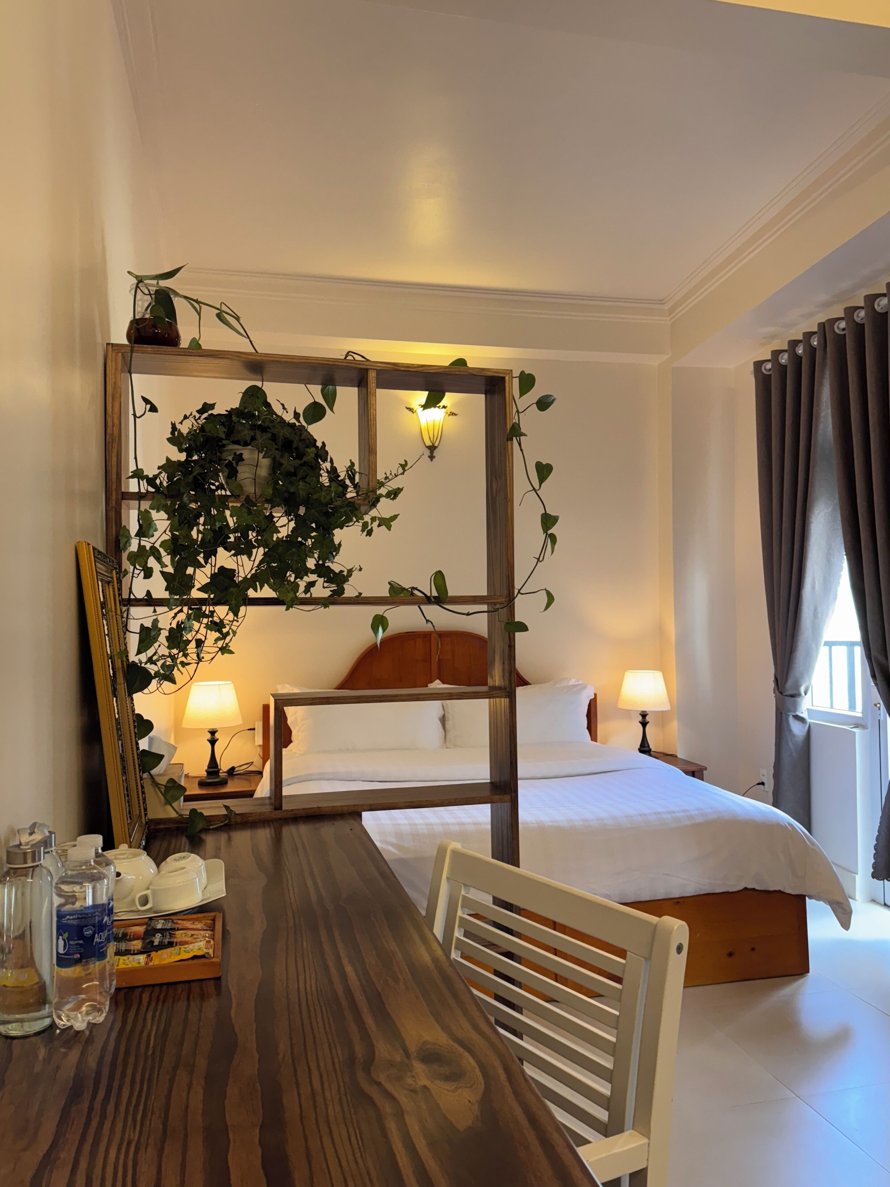 A hotel room with a wooden bed, two bedside lamps, a wooden table with a chair, a mirror, and a potted plant.