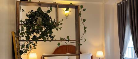 A hotel room with a wooden bed, two bedside lamps, a wooden table with a chair, a mirror, and a potted plant.